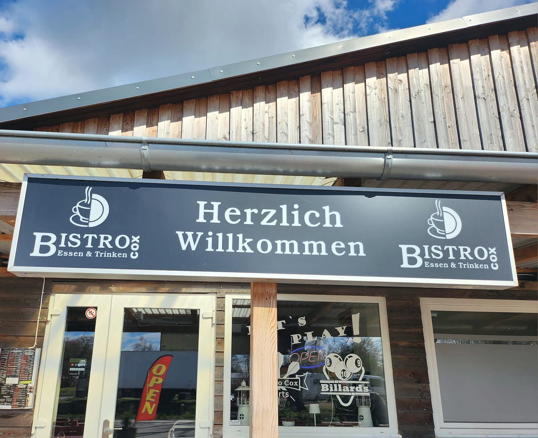 Außenansicht des Cox Bistro: Holzfassade, Leuchtschild «Herzlich Willkommen» mit Logo, Eingangstür und Fenster